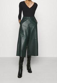 Schwarzes, geripptes Langarmoberteil kombiniert mit hoch taillierten, dunkelgrünen Leder-Culottes. Model trägt glänzende schwarze kniehohe Stiefel.