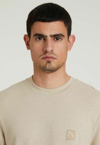 Bež dukserica s okruglim izrezom, koja ima mali logotip na gornjem lijevom dijelu. Tkanina izgleda mekano s glatkom teksturom.