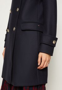 Gros plan d'un manteau bleu marine à double boutonnage avec des boutons dorés, des poches à rabat et un petit logo rectangulaire sur le rabat de la poche.