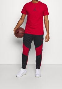 Rode T-shirt met korte mouwen en zwart logo, gecombineerd met zwarte en rode joggers met een geometrisch ontwerp. Houdt een basketbal vast. Witte sneakers.