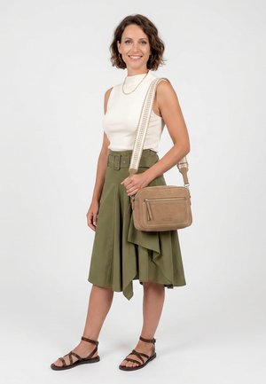 Kvinde iført hvid ærmeløs top, olivengrøn nederdel med bælte, brune sandaler, bærende en tan crossbody taske med mønstret strop, smilende og stående.
