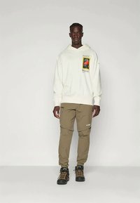 Ανοιχτόχρωμο φούτερ με γραφικά μπροστά, σε συνδυασμό με καμηλό tapered παντελόνι και μαύρες μπότες για πεζοπορία, μπροστά σε λευκό φόντο.