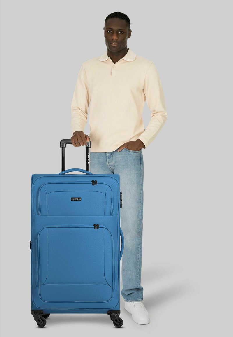 Homme en chemise beige à manches longues et jeans, debout, tenant la poignée d'une grande valise bleue à quatre roues sur fond gris.