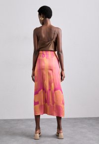 Top en satin marron avec des fines bretelles, associé à une jupe longue en dégradé de rose et d'orange. Modèle portant des sandales à talons beiges.