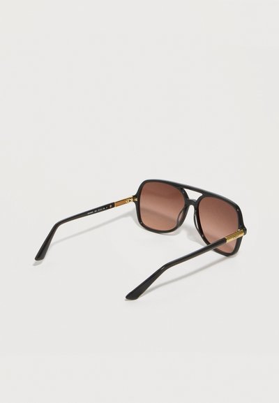 Μαύρα ορθογώνια γυαλιά ηλίου με χρυσές λεπτομέρειες και καφέ φακούς με απόχρωση, απεικονιζόμενα από πίσω σε λευκό φόντο σε ελαφριά γωνία.