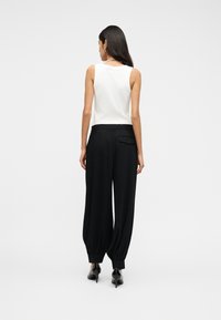 Γυναίκα με μακριά σκουρόχρωμα μαλλιά, φορώντας λευκό αμάνικο τοπ, μαύρο ψηλόμεσο φαρδύ παντελόνι με μανσέτες και μαύρα ψηλοτάκουνα παπούτσια, με την πλάτη στραμμένη.