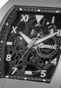 Montre mécanique avec un cadran squelette noir, des index des heures argentés, un boîtier en acier inoxydable et des engrenages visibles. Présente un mouvement automatique.