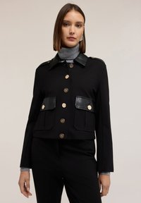 Giacca nera corta con bottoni dorati, due tasche frontali, colletto in pelle e dettagli sulle spalle. Tessuto liscio con un design strutturato.