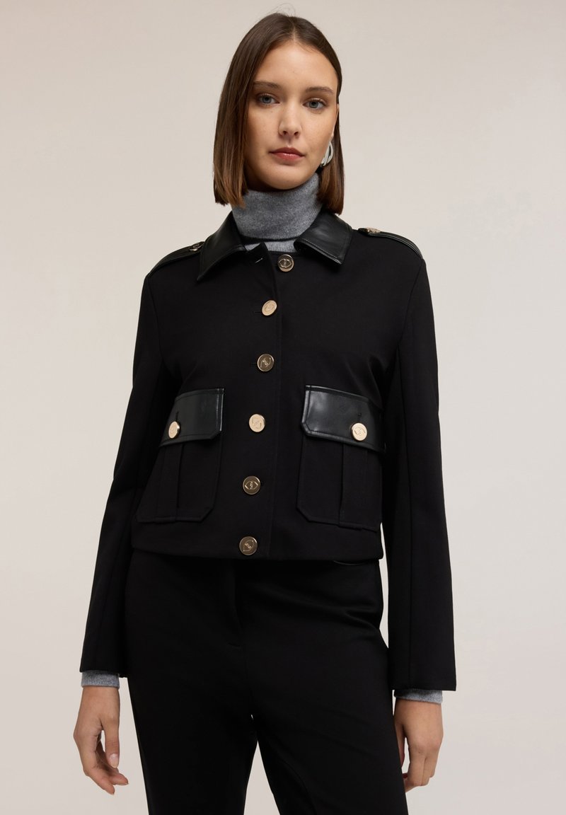 Giacca nera corta con bottoni dorati, due tasche frontali, colletto in pelle e dettagli sulle spalle. Tessuto liscio con un design strutturato.