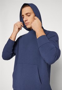 Muž upravuje kapuci na námořnicky modré mikině s kapucí, ukazuje malé tetování na zápěstí na obou pažích na jednobarevném pozadí.