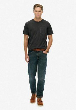 Young man standing, wearing a black t-shirt, blue jeans with rolled cuffs, brown belt, watch, and brown leather boots.
