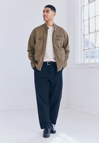 Bomber jakna olivno zelene barve z ovratnikom, dvema žepoma na prsih in gladko teksturo. Nošena čez svetlo srajco in ohlapne temno modre hlače.