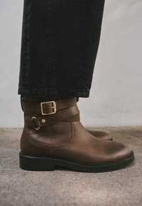 Bottes cheville en cuir marron à bout arrondi, dotées d'une double bride et d'une boucle en métal doré. Semelle en caoutchouc avec un talon bas.