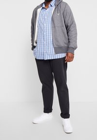 Haut à capuche gris zippé, chemise à carreaux bleus, pantalon noir et baskets blanches. Textures de tissu lisses ; le hoodie a des poches latérales et une doublure contrastante.