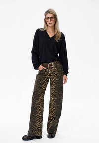 Schwarzes langärmeliges V-Ausschnitt-Oberteil kombiniert mit weit geschnittenen Leopardenmuster-Hosen. Mit einem braunen Gürtel mit silberner Schnalle und klobigen schwarzen Schuhen.