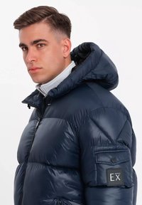 Giacca puffer blu navy con colletto alto e cappuccio. Presenta una chiusura con zip e una toppa sulla manica. Materiale liscio e lucido con design trapuntato.