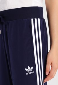 Mörkblå träningsbyxor med en bred elastisk midja, som har tre vita ränder längs sidorna och en broderad logotyp nära fickan.