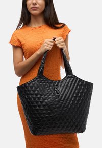 Tote bag mustavilla, jossa on kaksi tukevää kahvaa; etuosassa näkymätön logo. Valmistettu tekstuuritetusta nahkamateriaalista.