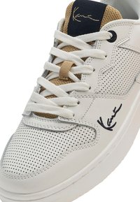 Zapatilla de cuero blanca con diseño perforado. Presenta una lengüeta de malla beis, cordones planos y un detalle característico en negro en el lateral.