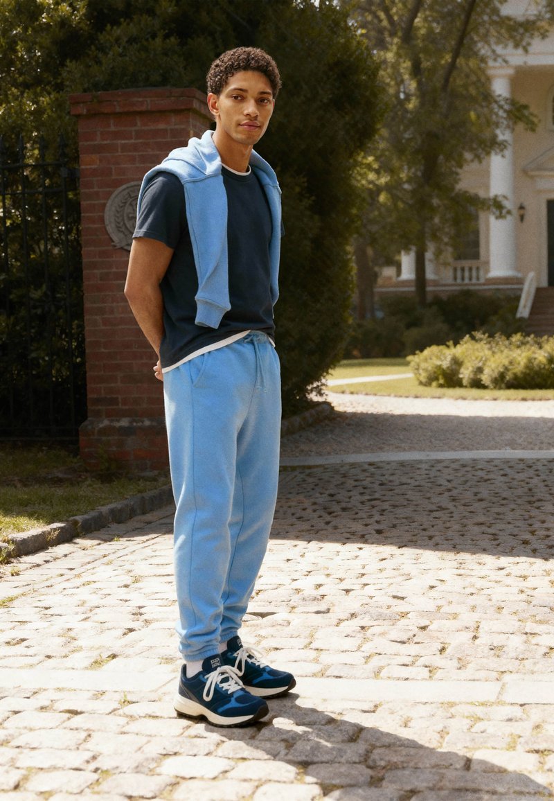 Jeune homme debout sur un chemin pavé près d'un pilier en briques, portant un pantalon de survêtement bleu, un t-shirt noir, un sweat-shirt bleu sur les épaules et des baskets bleues.