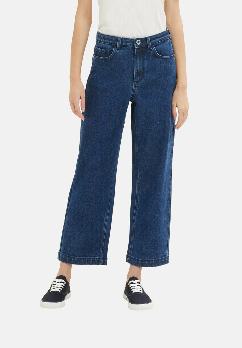 Personne portant un jean denim bleu foncé à jambes larges, un haut blanc et des baskets noires à lacets blancs, se tenant devant un fond uni.