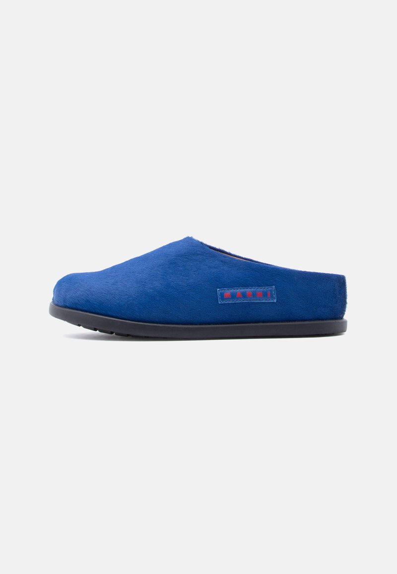 Chaussons bleu à enfiler en matériau doux et pelucheux avec une semelle en caoutchouc noir. Présentent une étiquette de logo rouge sur le côté.
