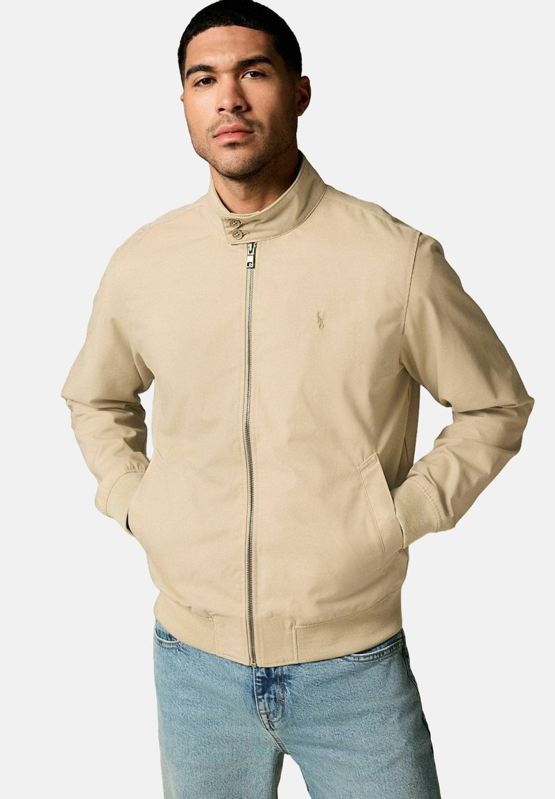 Veste zippée beige avec un col montant, des poignets élastiques et des poches latérales, fabriquée dans un tissu lisse arborant un petit logo sur la poitrine.