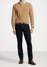 Suéter de punto trenzado en color beige claro, vaqueros oscuros, zapatos de cuero marrón. Camisa abotonada con rayas azul claro visible en el cuello.