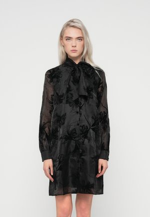 Sort gennemsigtig kjole med blomsterbroderi, der har en høj krave og sløjfe detalje. Lange ærmer og knaplukning fuldender designet.