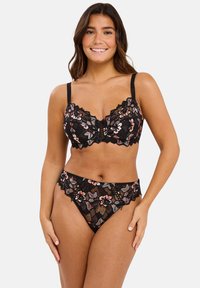 Femme souriante portant un soutien-gorge en dentelle noire et un shorty taille haute en dentelle assorti, ornés de broderies florales sur un fond gris clair.