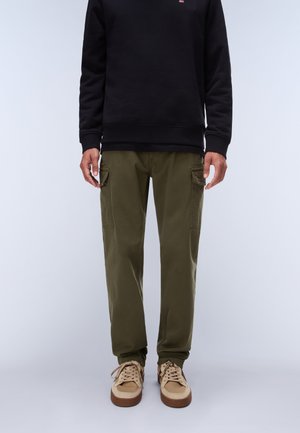 Uomo con pantaloni cargo verde oliva, sneakers beige e maglia nera a maniche lunghe in piedi davanti a uno sfondo chiaro e uniforme.