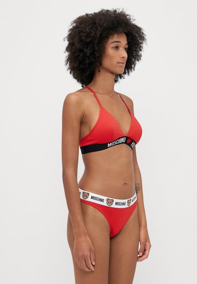 Sarkans bikini komplekts ar trijstūra veida krūšturi un atbilstošām biksītēm, ar melnu joslu, uz kuras ir rakstīts "Moschino" un lāču ikona uz baltas elastīgas auduma.