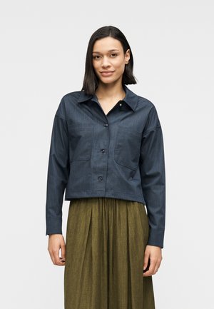 Dunkelblaue Jacke mit Kragen und Knopfleiste, zwei Brusttaschen und langen Ärmeln, kombiniert mit einem plissierten olivgrünen Rock.