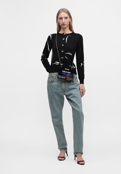 Femme portant un cardigan noir à motifs, un jean bleu clair, des sandales noires à talons hauts et un petit sac à bandoulière au design de bande cassette.