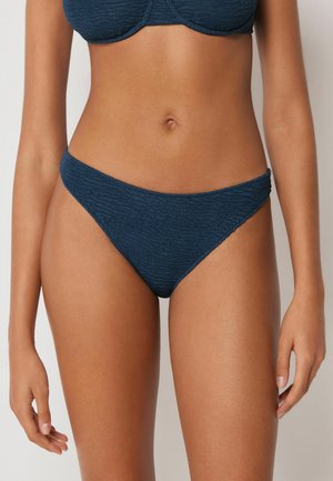 Frau trägt einen strukturierten dunkelblauen Bikinihose und passenden Oberteil, steht mit locker an den Seiten hängenden Händen vor einem einfarbigen Hintergrund.