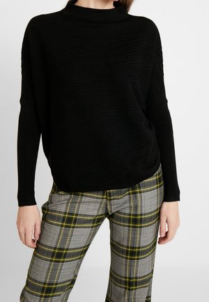 Maglione nero a maglia con colletto alto e motivo a onde testurizzato, abbinato a pantaloni a quadri grigi con motivi gialli e verdi.