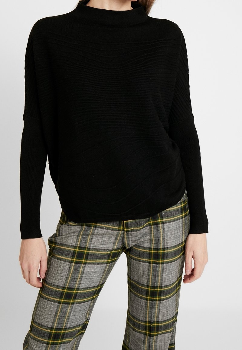 Svart stickad tröja med hög krage och strukturerat vågmönster, i kombination med grå rutiga byxor med gula och gröna rutor.