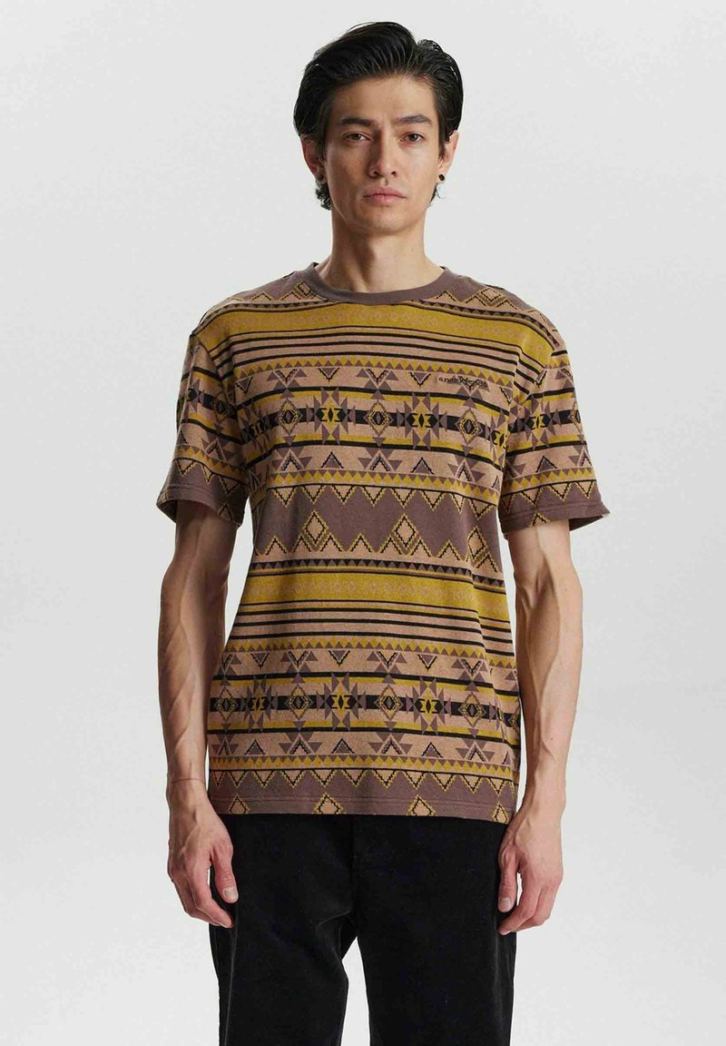Camiseta de manga corta, de color marrón, con un patrón que presenta diseños geométricos y tribales en amarillo y negro, fabricada en un tejido texturizado.