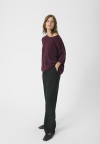 Burgundowy, przerośnięty sweter z dzianiny z rękawami trzy czwarte, zestawiony z czarnymi, dopasowanymi spodniami oraz płaskimi czarnymi butami, stojący na tle białym.