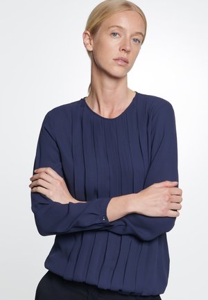 Blusa blu navy a maniche lunghe con pieghe verticali, scollatura rotonda e polsini con dettagli di bottoni. Tessuto liscio con una texture morbida.