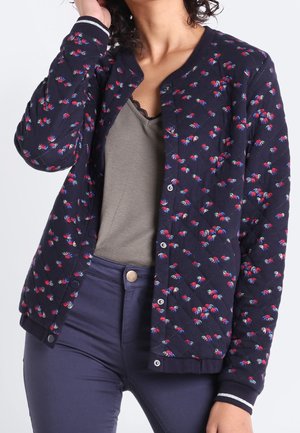 Frau trägt eine marineblaue gesteppte Jacke mit kleinen roten und blauen Blumenmustern über einem grauen Oberteil und dunkelblauen Jeans, während sie mit einer Hand ihre Haare richtet.