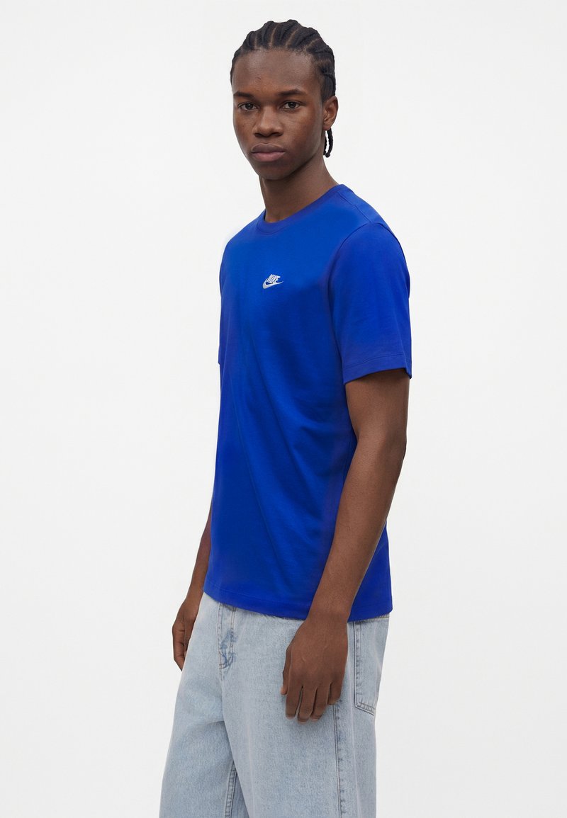 Junger Mann mit geflochtenem Haar, der ein königsblaues Nike T-Shirt und hellblaue Jeans trägt, steht vor einem schlichten weißen Hintergrund.