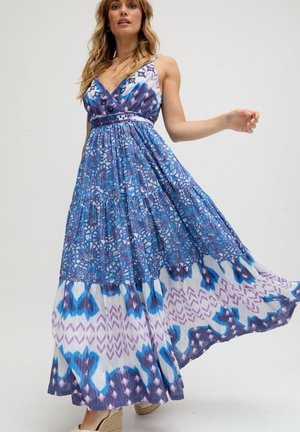 Frau trägt ein fließendes, ärmelloses Maxikleid mit blauem, lila und weißem Muster, steht auf einem Fuß und hebt dabei den Rock an.