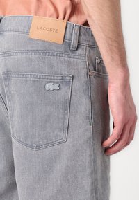 Grå denimjeans med et lærlogo fra Lacoste, har en baklomme med en brodert krokodillelogo og subtil tekstur.