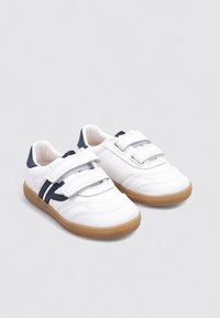 Zapatillas deportivas blancas con acentos en azul marino, que cuentan con correas de velcro, un material suave similar al cuero y una suela de goma marrón.