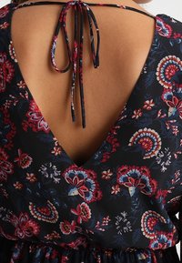 Blumenmusterkleid mit tiefem V-Ausschnitt auf der Rückseite, strukturiertem schwarzen Stoff mit roten und blauen Blumen sowie einer Schleifenbindung am Ausschnitt.