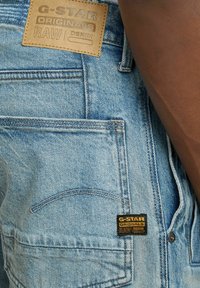 Vaaleansinisiä farkkuhousuja, joissa on teksturoitu pinta. Housuissa on vaaleanruskea nahkapäällyste ja musta etiketti, jossa on brändin nimi ja tyylitiedot.