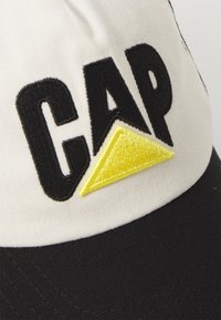 Valge ja musta kangast baseballi müts, mille esiosal on musta värvi broderdatud "CAP" ning kollane kolmnurkne aksent.