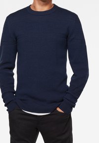 Marineblauer Strickpullover mit strukturiertem Muster über Brust und Ärmeln, rundem Halsausschnitt sowie gerippten Bündchen und Saum. Trägt dazu schwarze Hose.