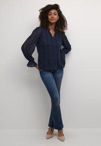 Blusa di pizzo blu navy con scollo a V e maniche lunghe, caratterizzata da motivi geometrici, abbinata a jeans blu aderenti e tacchi beige.